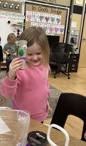 School Age 1 child standing holding a tube with google eyes, a pom pom nose, and ribbon around it - School Age child standing in the back