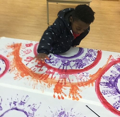 child at the table adding an orange handprint to a painting of a rainbow