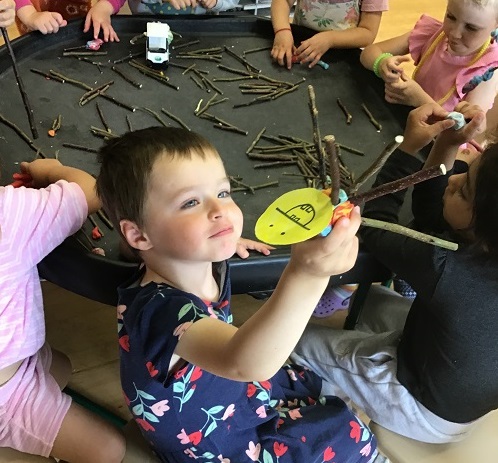 child sitting around an activity table full of twigs and sticks. They are holding up a smiley face drawn onto yellow paper that is attached to a bundle of sticks. 