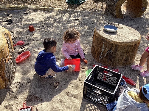 2 children exploring with shovels, scoops, bowls, colanders and other materials in the sandbox