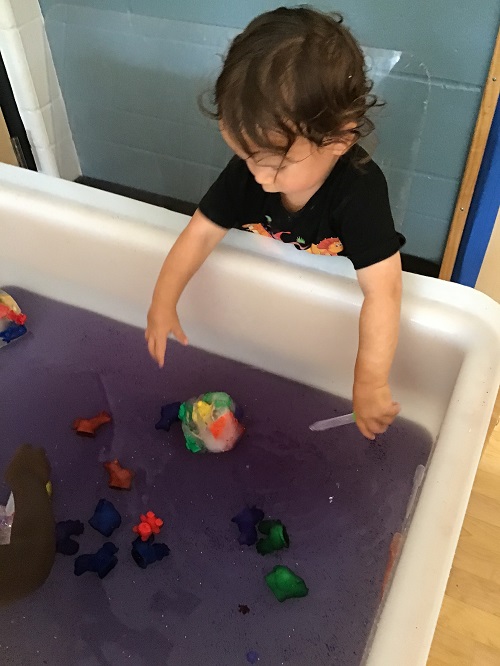 child standing with their arms hanging inside of a large water table with purple water and different coloured animals floating in the water