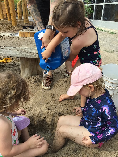 two childing sitting with their feet in a hole full of water in the sandbox while another child is pouring more water into the hole from a large blue jug