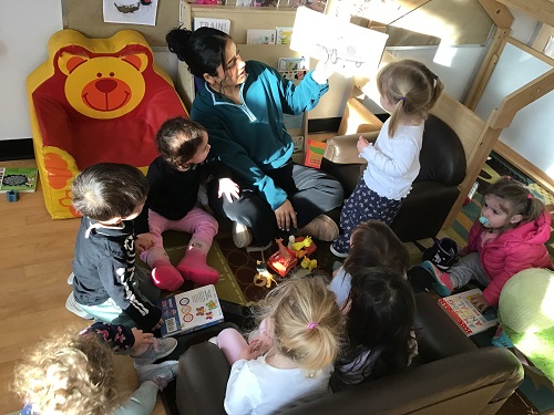 8 toddler children sitting on the carpet and couches and looking at a story book that the educator is holding up for the children