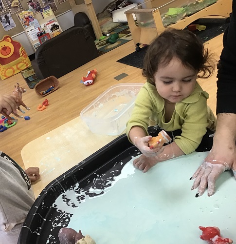 toddler child standing at the sensory table filled with oobleck holding a toy animal over the oobleck