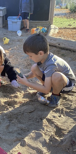 2 School age children in the sandbox, one has their hand in the sand, the other is holding and looking at a dinosaur in their hand