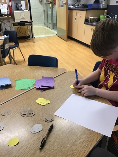 School Age child sitting at the table writing money amounts on cut out construction paper