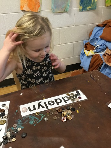 A child sitting at a table placing buttons on the letters of the name card in front of her