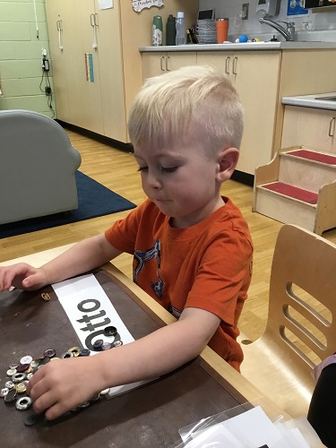 A child sitting at a table placing buttons on the letters of the name card in front of him