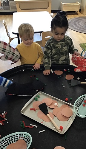 Two children standing at the tuff tray with oven mitts on, looking at the materials in front of them