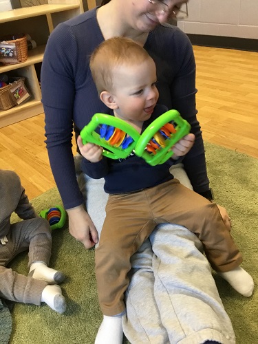 A child sitting on an educator's lap holding shakers in both hands