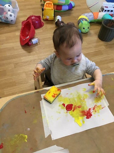 An infant sitting at a table with a sponge and paper covered in paint in front of him