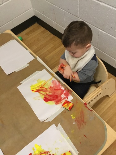 An infant sitting at a table rubbing paint on his arms