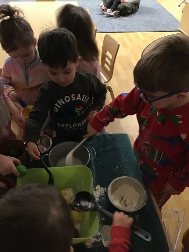 Children standing beside a table using spoons to mix play food in a large bowl