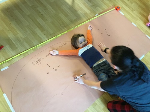 A child laying down on a paper with outstretched arms while an educator measures him