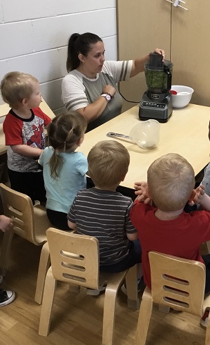 Children sitting around a table watching an educator turn on a food processor