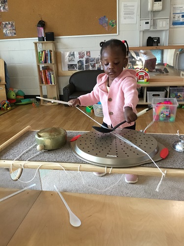 A child using a large spoon to bang on a shelf covered in metal pots and pans