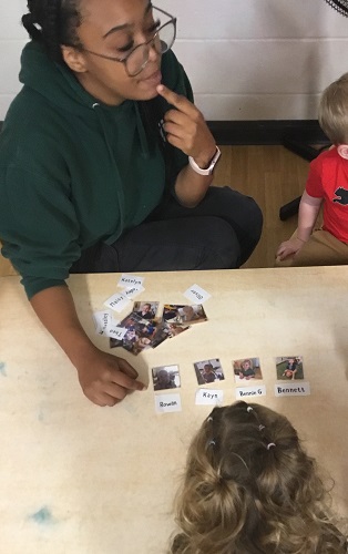 An educator at a table with a group of children, pointing to her mouth as she models how to say different names