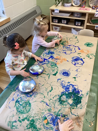 Two children using blocks to paint on a long brown piece of paper