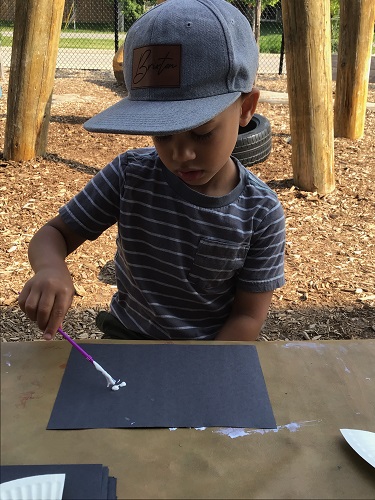 A child sitting at a table using a paint brush to spread sunscreen on a piece of black construction paper