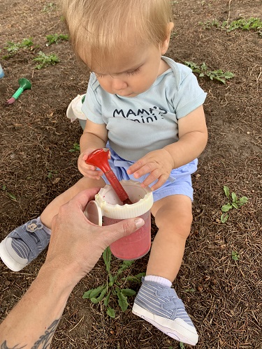 A child sitting on the ground dipping a paint brush into a paint pot 