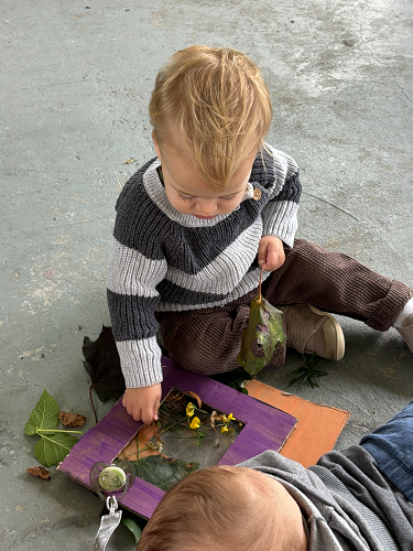 A child sitting on the ground sticking leaves and flowers to a cardboard frame