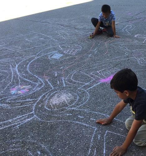Children sitting on the blacktop driving toy cars on chalk drawn roads 