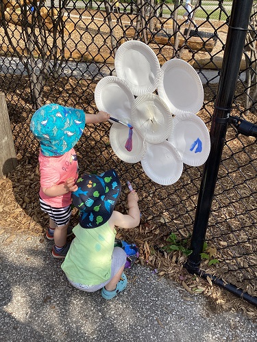 Children outside at the fence painting a paper plate flower