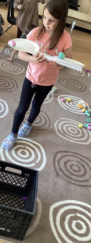 School age child holding a cardboard tube with paper plates attached to the ends with balls balancing ontop of the plates