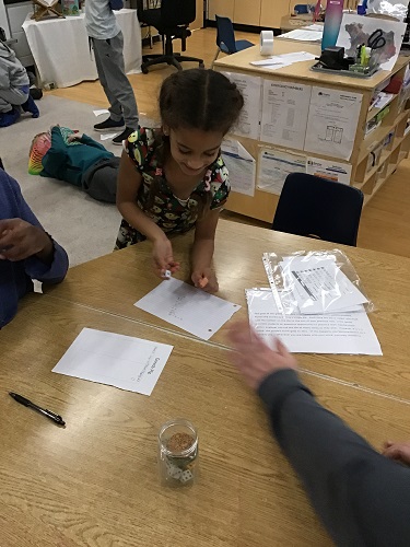 School Age 2 child standing at the table holding a die in her hand and smiling, another School Age 2 child laying on the floor in the background with a slinky over his face and another School Age 2 child standing in the background