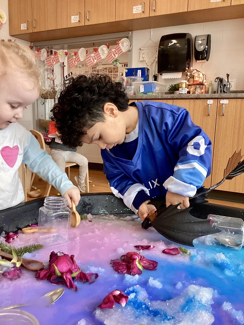 2 preschool children working at a tuff tray with snow, ice, spoons, small containers and flowers