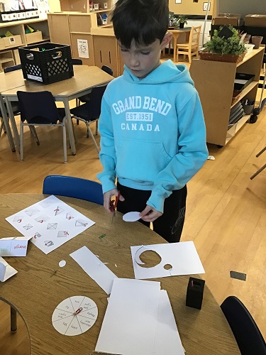 1 School Age child standing at the table holding scissors and a round piece of paper looking at the instructions on the table