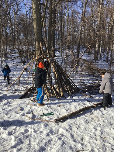 3 School Age children in the photo, one school age children putting a log up on the tent, another school age child looking down at a log on the ground and another school age child walking towards the tent.