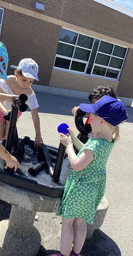 4 children outside standing around a tuff tray with water and tubes and small cups