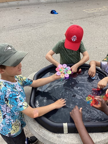4 children standing around a tuff tray with water and connectors, one child is looking at another child beside them