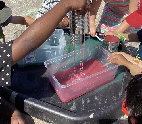 5 children standing around watching another child hold up a tin and watching the colourful water pour out of holes in the bottom