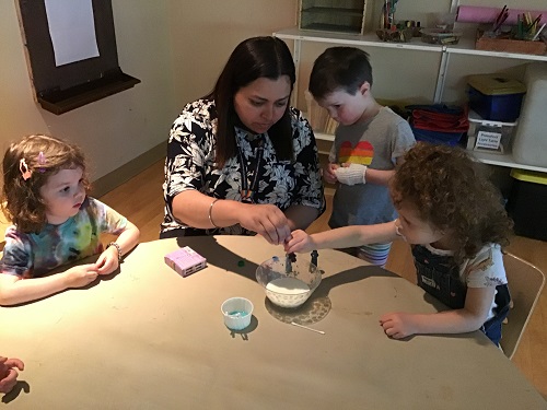An educator, along with several preschool children, are using droppers to add food colouring to milk.