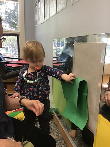 An infant is standing in front of a mirror and holding a piece if paper onto the mirror so an educator can tape it up.