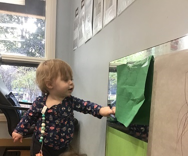 An infant is colouring on a piect of paper that is taped to a mirror.