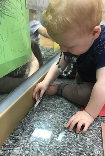 An infant is sitting in front of a mirror and picking up a crayon they have dropped while colouring.