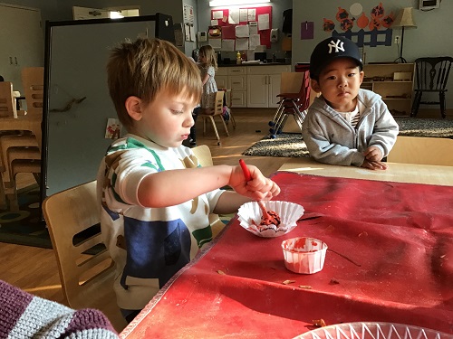 A preschooler is painting a coffee filter as another preschooler watches.