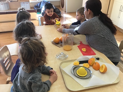 A group of preschoolers are sitting at a table, taking turns squeezing the juice from an orange, with different tools.