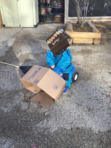 A toddler is riding on a car with a box on the front, and a box on their head.