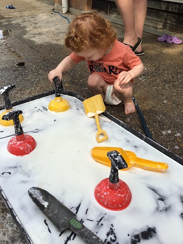 A toddler is using a small plunger to play in Oobleck.