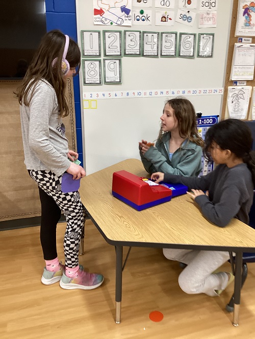 2 school age children sitting at a table with a cash register with another school age child standing behind the table