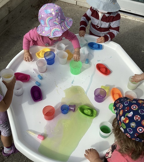 A group of children exploring with coloured water and various items in the white tuff tray.