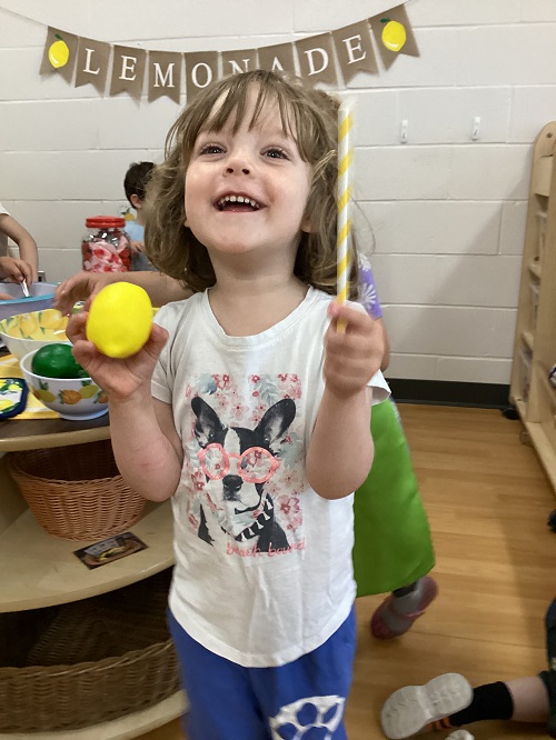 A child happily holding up items from the dramatic play lemonade stand.