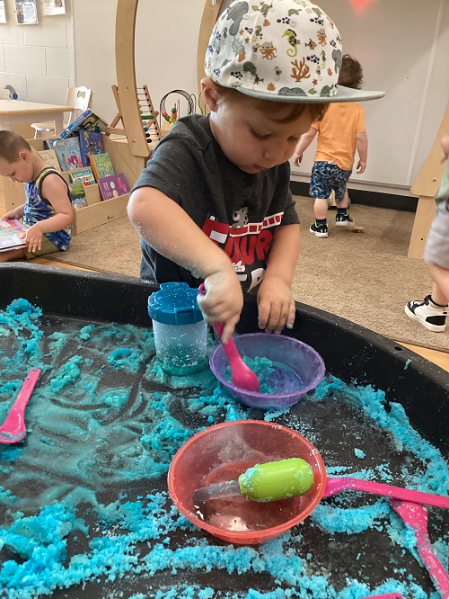 A child using various materials to explore with coloured salt in a tuff tray.