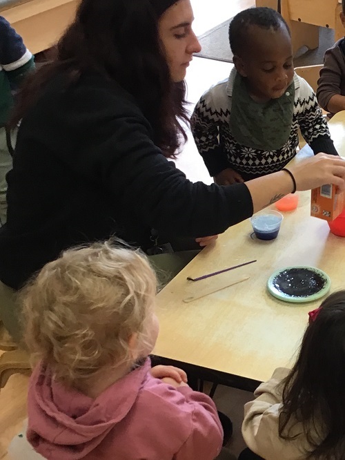 4 Toddler children sitting at the table watching, one Toddler is standing up and reaching towards the baking soda the educator is pouring into a separate container