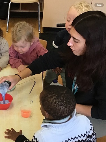 3 Toddler children sitting at the table and one is standing, they are all looking over the educator to watch as she pours purple liquid into a bowl. 