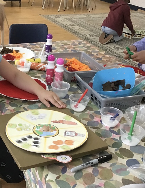 School Age child sitting on the carpet in the background. Child sitting at the table with a spinner of vegetables, and felt vegetables in front of them on the table